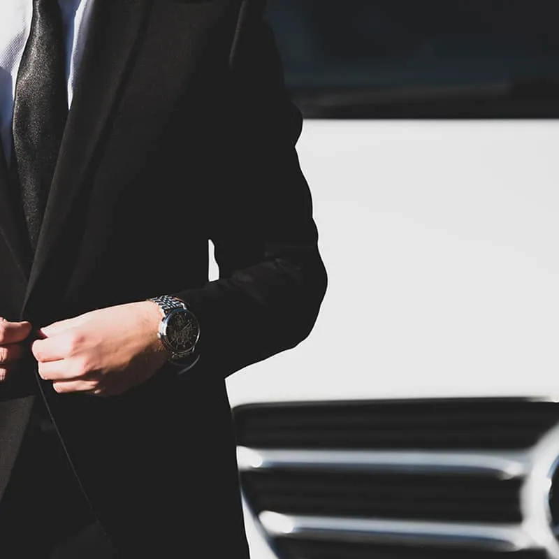 Homme en costume noir avec une montre devant une voiture