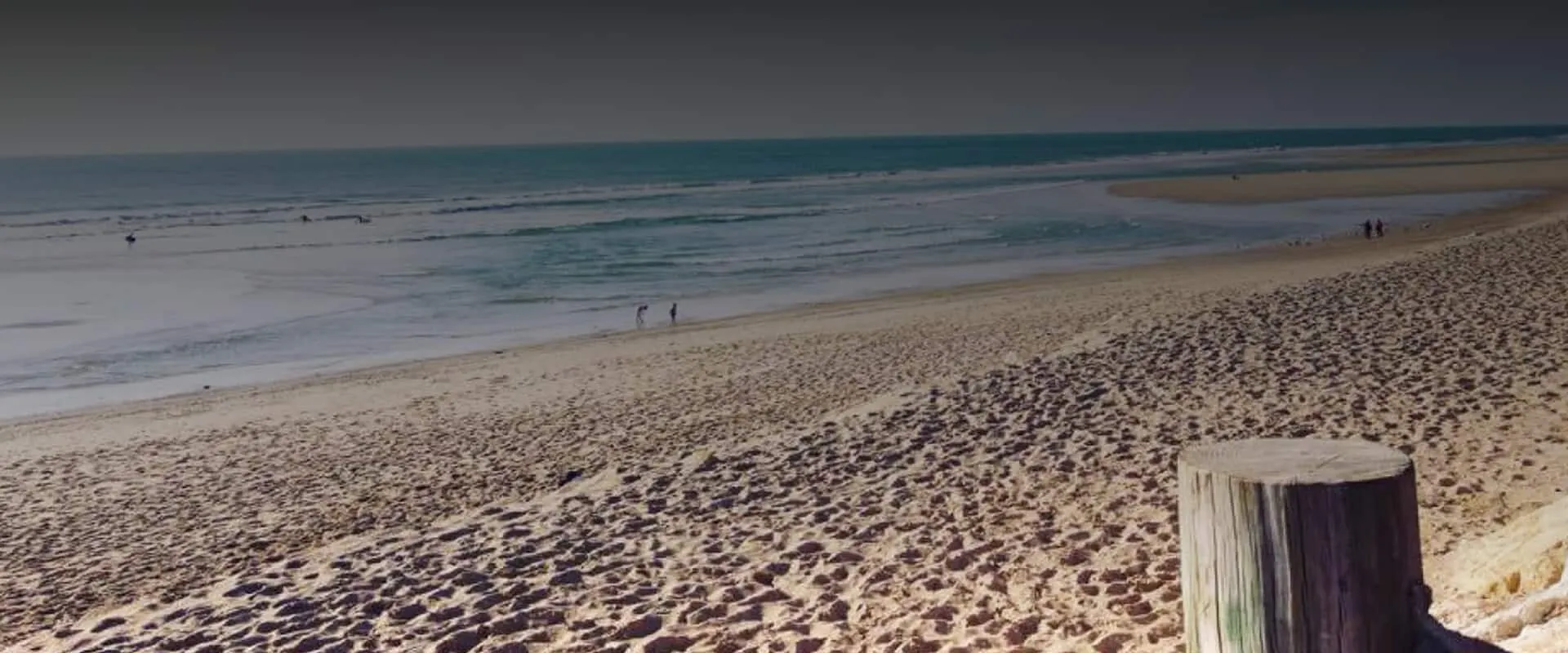 Plage de sable en bord d'océan avec des promeneurs et un pieu en bois
