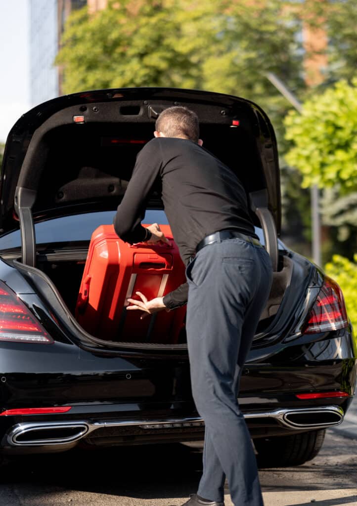 Chargement valise rouge dans coffre voiture noire Homme en chemise noire et pantalon bleu charge une valise rouge dans le coffre ouvert d'une berline noire luxueuse.