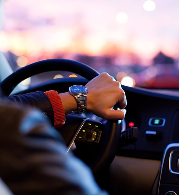 Conducteur à la montre de luxe au coucher du soleil Gros plan sur la main d'un conducteur portant une montre-bracelet argentée, tenant le volant au crépuscule avec des lumières de ville floues en arrière-plan.