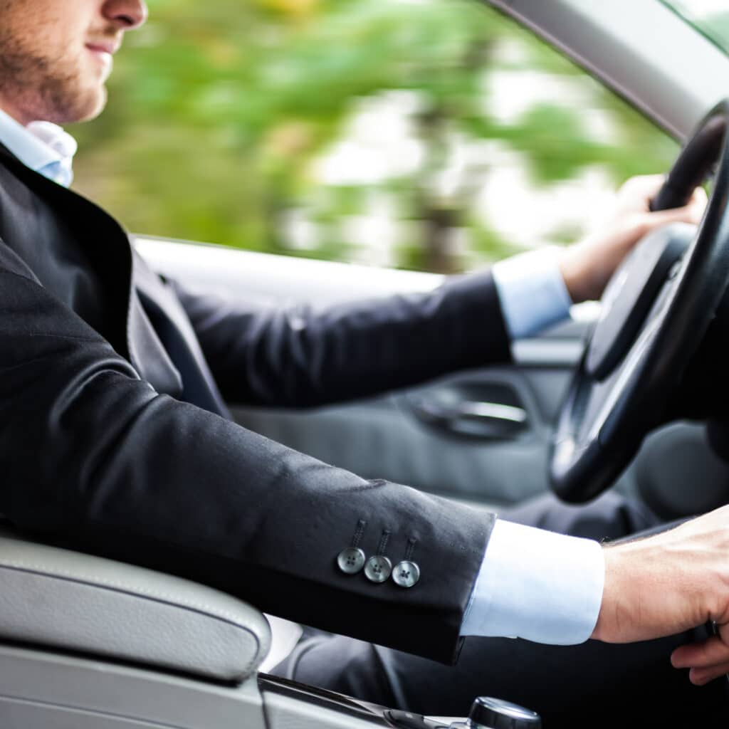 Homme d'affaires conduisant une voiture en vitesse Homme d'affaires en costume noir conduisant, vue rapprochée sur la manche à boutons et les mains sur le volant, arrière-plan flou de verdure.