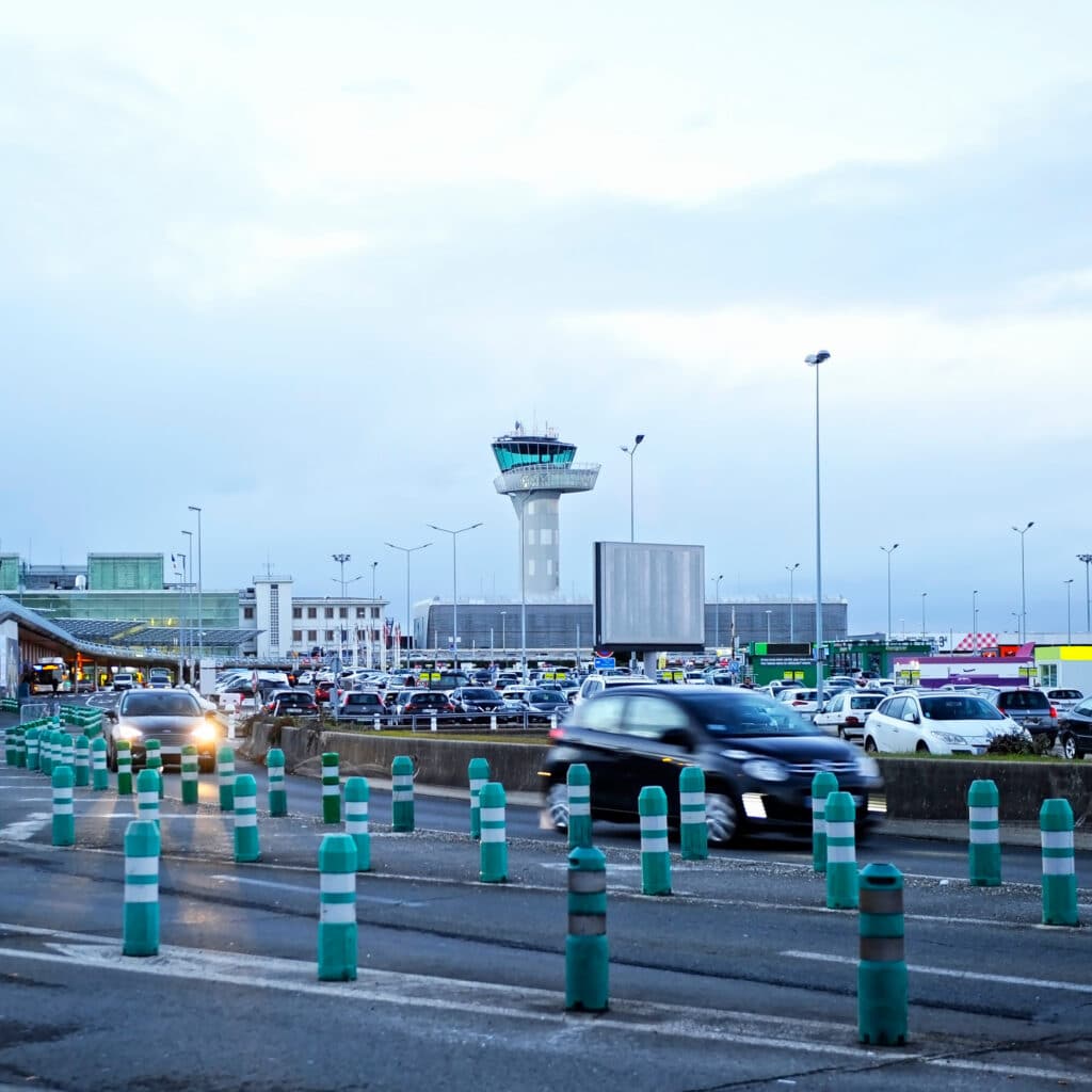 Tour de contrôle aéroportuaire et trafic, arrivée voyageurs Aéroport avec tour de contrôle, trafic automobile dense et bornes vertes en premier plan, ciel gris-bleu.