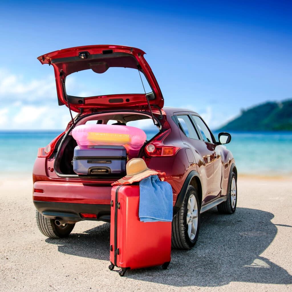 Valises prêtes pour voyage en voiture vers la plage Voiture rouge garée près de la mer, coffre ouvert chargé de valises et d'une bouée rose. Bagages prêts pour les vacances.