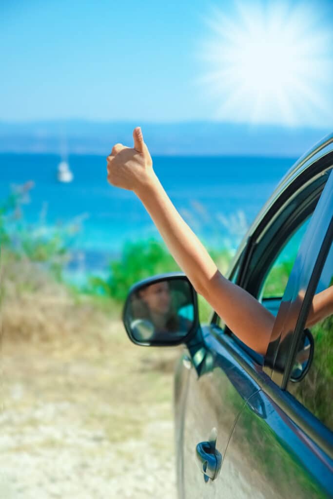 Vacances en voiture : pouce levé bord de mer ensoleillé Main faisant un pouce levé sortant de la fenêtre d'une voiture avec une mer turquoise et un soleil éclatant en arrière-plan.